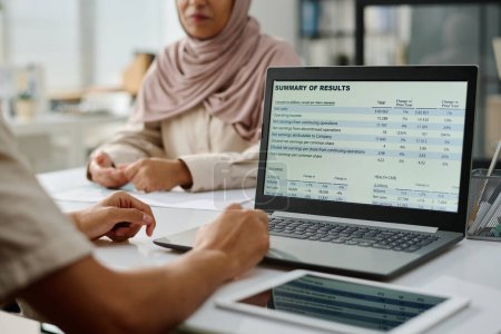 Téléchargez les photos : Informations financières à l'écran de l'ordinateur portable utilisé par un jeune homme d'affaires musulman assis devant une collègue féminine dans le hijab - en image libre de droit