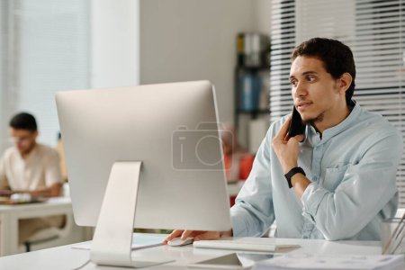 Téléchargez les photos : Jeune homme gestionnaire confiant tenant smartphone par l'oreille alors qu'il était assis devant l'ordinateur de bureau dans le bureau et téléphone collègue - en image libre de droit