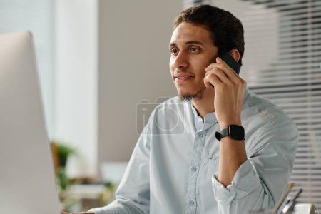Téléchargez les photos : Jeune employé masculin parlant à quelqu'un par téléphone portable alors qu'il était assis devant un ordinateur de bureau et faisant défiler les données en ligne - en image libre de droit