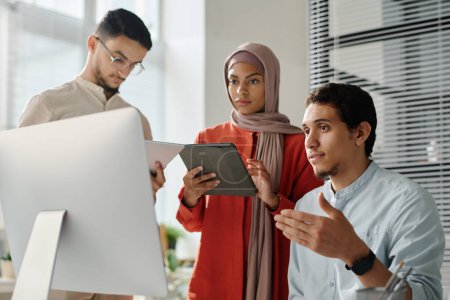 Téléchargez les photos : Deux jeunes employés musulmans utilisant des tablettes alors qu'ils se tenaient à côté de leur collègue regardant l'écran d'ordinateur et expliquant les données en ligne - en image libre de droit