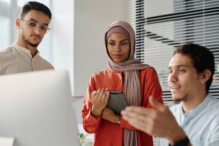 Photo for Young businesswoman in hijab with tablet in hands looking at computer screen and listening to explanation of data of male colleague - Royalty Free Image
