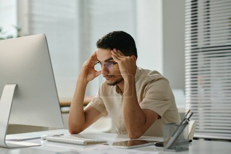 Téléchargez les photos : Jeune employé masculin stressé assis par bureau devant l'écran de l'ordinateur et attendant le téléchargement de la page tout en essayant de se concentrer - en image libre de droit
