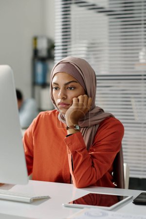 Téléchargez les photos : Jeune femme d'affaires ennuyée dans le hijab assis devant l'ordinateur de bureau et regardant l'écran en attendant le téléchargement de la page - en image libre de droit