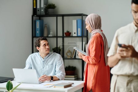 Photo for Young businessman with laptop sitting by workplace and looking at female colleague in hijab using tablet and saying something to man - Royalty Free Image