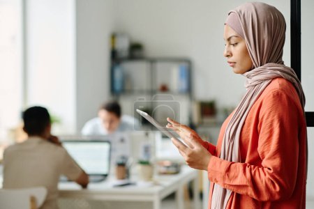 Téléchargez les photos : Jeune employée musulmane dans le hijab utilisant une tablette tout en se tenant devant la caméra dans un espace de coworking avec des collègues masculins - en image libre de droit