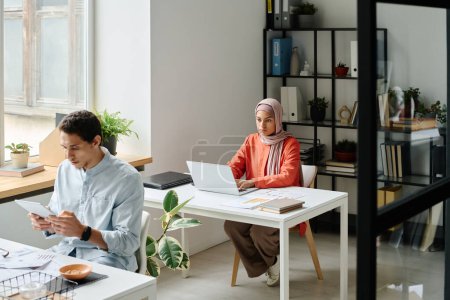 Téléchargez les photos : Jeune femme d'affaires musulmane dans le hijab assis par le lieu de travail dans l'espace de coworking et regardant l'écran d'ordinateur portable tout en analysant les données - en image libre de droit