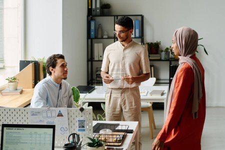 Photo for Group of three young intercultural Muslim business people discussing points of new project at start-up meeting in coworking space - Royalty Free Image