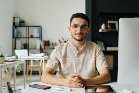 Téléchargez les photos : Jeune homme musulman PDG ou entrepreneur assis sur le lieu de travail avec ordinateur de bureau dans l'espace de coworking et en regardant la caméra - en image libre de droit