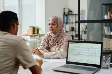 Téléchargez les photos : Jeune femme d'affaires musulmane confiante dans le hijab expliquant son point de vue à un collègue masculin et le regardant lors d'une réunion par lieu de travail - en image libre de droit