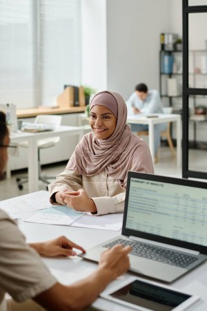 Téléchargez les photos : Jeune économiste musulmane souriante en hijab regardant un collègue masculin à l'aide d'un ordinateur portable tout en analysant les données financières à l'écran - en image libre de droit