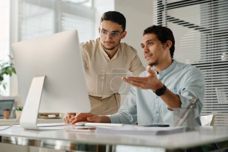 Foto de Dos jóvenes empresarios musulmanes mirando la pantalla de la computadora de escritorio, mientras que uno de ellos explica los datos en línea a su colega - Imagen libre de derechos