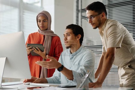 Photo for Team of three young Muslim employees looking at computer screen while confident man explaining online data to his colleagues - Royalty Free Image