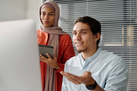 Téléchargez les photos : Jeune analyste musulman confiant expliquant les données à l'écran de l'ordinateur à une collègue féminine dans le hijab lors de la présentation des points du projet - en image libre de droit