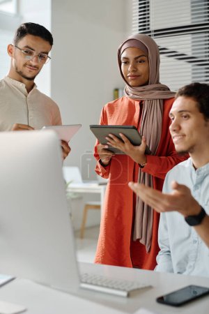 Téléchargez les photos : Deux jeunes courtiers musulmans avec des tablettes regardant l'écran de l'ordinateur tout en écoutant un collègue masculin leur expliquer les données en ligne - en image libre de droit