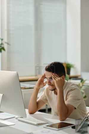 Téléchargez les photos : Travailleur de bureau masculin anxieux essayant de se concentrer tout en étant assis devant l'écran d'ordinateur et en regardant la page de téléchargement - en image libre de droit