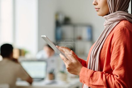 Téléchargez les photos : Plan recadré de jeunes femmes musulmanes gestionnaires utilisant une tablette tout en se tenant dans l'espace de coworking contre des collègues masculins - en image libre de droit