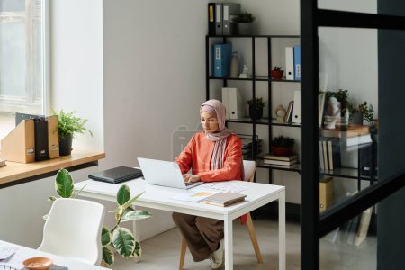 Téléchargez les photos : Jeune analyste musulmane dans le hijab assise par bureau dans l'espace de coworking, regardant l'écran de l'ordinateur portable et analysant les données en ligne - en image libre de droit