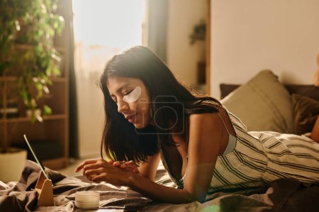 Foto de Mujer joven relajándose en la cama en el ambiente hogareño y disfrutando de la mañana de fin de semana mientras aplica crema en sus manos - Imagen libre de derechos