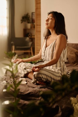 Téléchargez les photos : Jeune femme latine sereine en pyjama assis sur le lit en pose de lotus le matin le week-end et profiter de la pratique du yoga - en image libre de droit