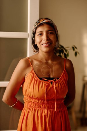 Foto de Mujer sonriente joven en ropa de casualwear inteligente mirando a la cámara con sonrisa mientras está de pie contra la puerta abierta de la sala de estar de su apartamento - Imagen libre de derechos