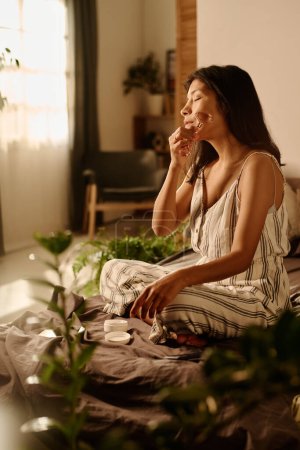 Foto de Joven mujer complacida con los ojos cerrados disfrutando de masaje facial con rodillo de piedra de jade mientras se sienta en la cama por la mañana durante el procedimiento - Imagen libre de derechos