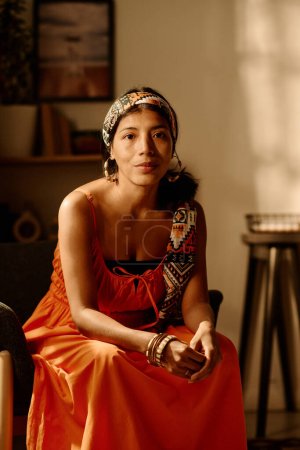 Foto de Joven mujer hispana serena en pañuelo étnico y vestido rojo mirando a la cámara mientras está sentada en un sillón en la sala de estar del apartamento moderno - Imagen libre de derechos