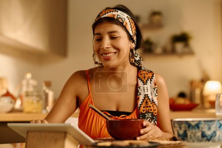 Téléchargez les photos : Jeune femme hispanique souriante dans le bandeau regardant l'écran de la tablette alors qu'elle était assise à table dans la cuisine, prenant le petit déjeuner et regardant la vidéo - en image libre de droit