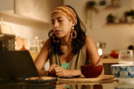 Téléchargez les photos : Jeune femme d'affaires élégante en tenue de maison regardant l'écran d'ordinateur portable tout en étant assis à la table de cuisine après le petit déjeuner et en travaillant sur un nouveau projet - en image libre de droit