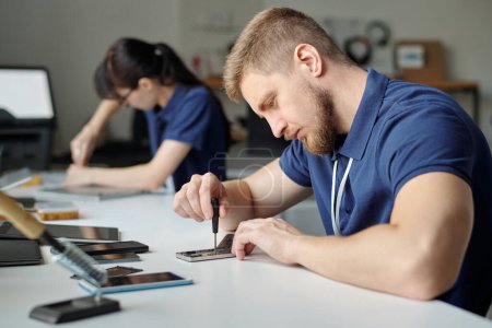 Photo for Young male technician or repairman bending over desk and fixing tiny details of smartphone with screwdriver against female colleague - Royalty Free Image