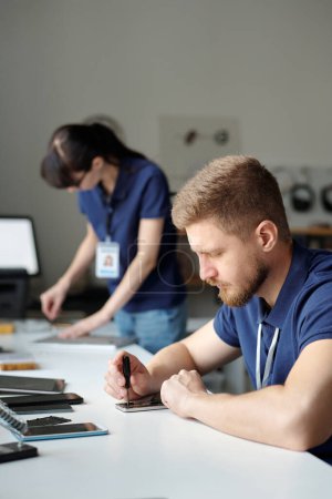 Photo for Young technician sitting by workplace in repair center office and fixing tiny details of broken mobile phone with screwdriver against colleague - Royalty Free Image