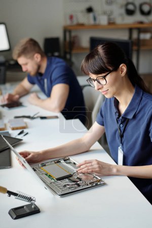 Photo for Young brunette technical service engineer checking details of disassembled laptop while sitting by desk against male colleague - Royalty Free Image