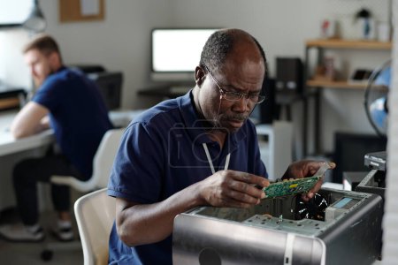 Photo for Experienced African American repairman in eyeglasses looking at computer motherboard and identifying technical problems in office - Royalty Free Image