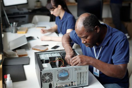 Téléchargez les photos : Technicien professionnel assis par bureau dans le bureau de service de réparation et se penchant sur le processeur d'ordinateur démonté tout en le vérifiant - en image libre de droit