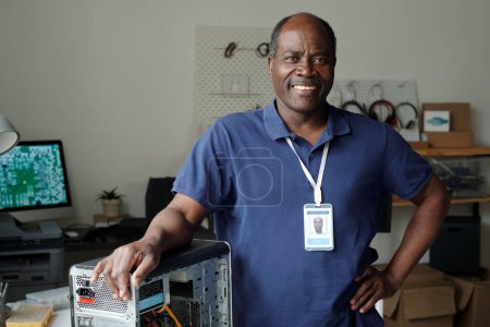 Reifer afroamerikanischer Techniker in blauem Hemd, der den Arm über dem Computerprozessor hält und am Arbeitsplatz in die Kamera blickt