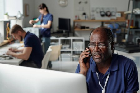 Téléchargez les photos : Technicien masculin mature du service de réparation assis par le lieu de travail devant l'écran d'ordinateur et parlant au client sur le téléphone mobile - en image libre de droit