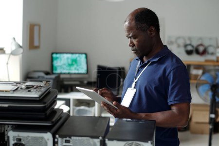 Foto de Técnico afroamericano maduro serio con la tableta parada delante de la cámara por el lugar de trabajo y mirando a través del manual en línea - Imagen libre de derechos