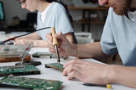 Photo for Young technical specialist of maintenance service center sitting by workplace and repairing computer motherboard with electric handtool - Royalty Free Image