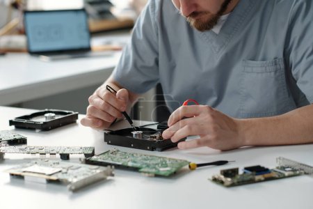 Photo for Cropped shot of young man repairing detail of computer processor with electric transmission tweezers while sitting by workplace - Royalty Free Image