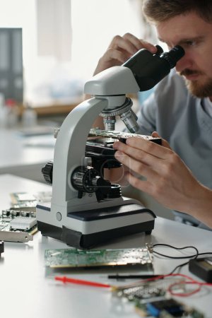 Téléchargez les photos : Microscope et main du jeune technicien du centre de service de réparation regardant les détails de la carte mère assis devant la caméra en laboratoire - en image libre de droit