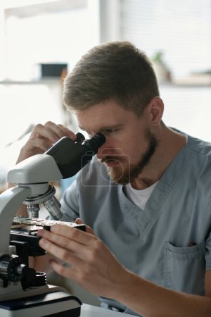 Téléchargez les photos : Jeune technicien masculin sérieux du centre de dépannage regardant la carte mère au microscope tout en étant assis par le lieu de travail - en image libre de droit