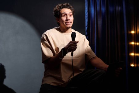 Junger männlicher Stand-up-Comedian, der seinen Monolog vorträgt, während er auf einem Hocker auf der Bühne des Konzerthauses sitzt und ins Mikrofon spricht