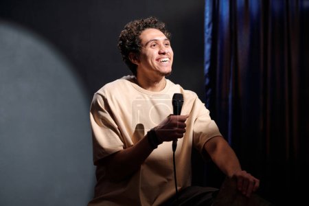 Joven comediante masculino alegre con micrófono en la mano realizando un nuevo monólogo frente a la audiencia mientras está sentado en el escenario de la sala de conciertos