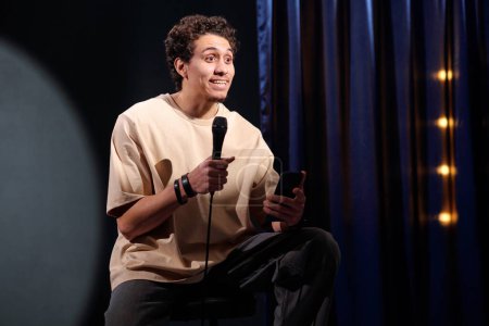 Young comedian with microphone and mobile phone in hands performing monologue in front of audience on stage of stand up club