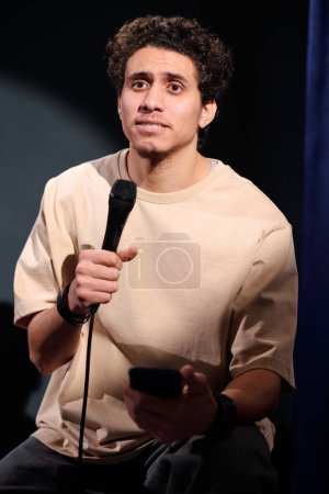 Téléchargez les photos : Jeune comédien de stand up club parlant au microphone et utilisant smartphone pendant le monologue devant le public sur scène - en image libre de droit