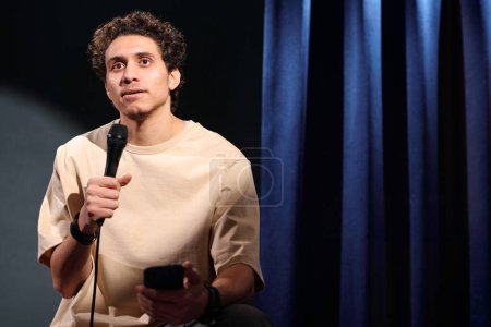 Joven comediante masculino con micrófono en la mano usando smartphone durante el monólogo mientras actúa en el escenario frente a la audiencia