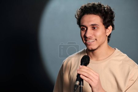 Téléchargez les photos : Jeune homme souriant parlant au microphone et regardant le public tout en se tenant sur scène du stand up club pendant le monologue - en image libre de droit