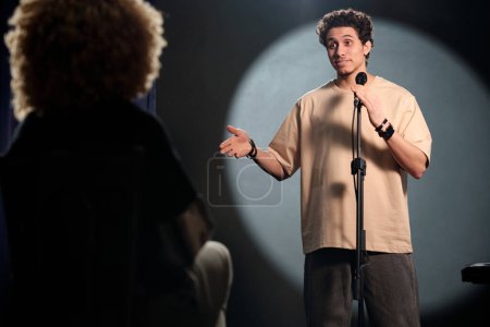 Photo for Young man in t-shirt speaking in microphone and looking at audience or group of judges during rehearsal or stand up performance - Royalty Free Image