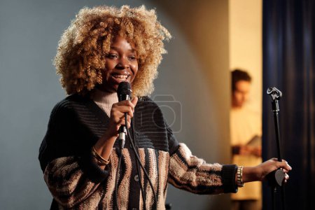 Fröhliche junge Schauspielerin oder Komikerin spricht ins Mikrofon, während sie auf der Bühne vor Publikum steht und einen neuen Monolog vorträgt