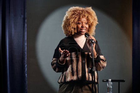 Téléchargez les photos : Jeune afro-américain debout comédien en perruque parlant au microphone pendant la performance tout en se tenant sur scène devant le public - en image libre de droit