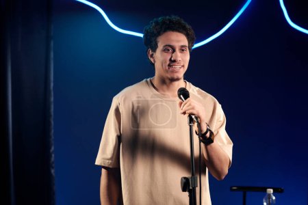 Téléchargez les photos : Jeune comédien homme en t-shirt beige parlant au microphone sur scène en regardant le public tout en prononçant un monologue - en image libre de droit